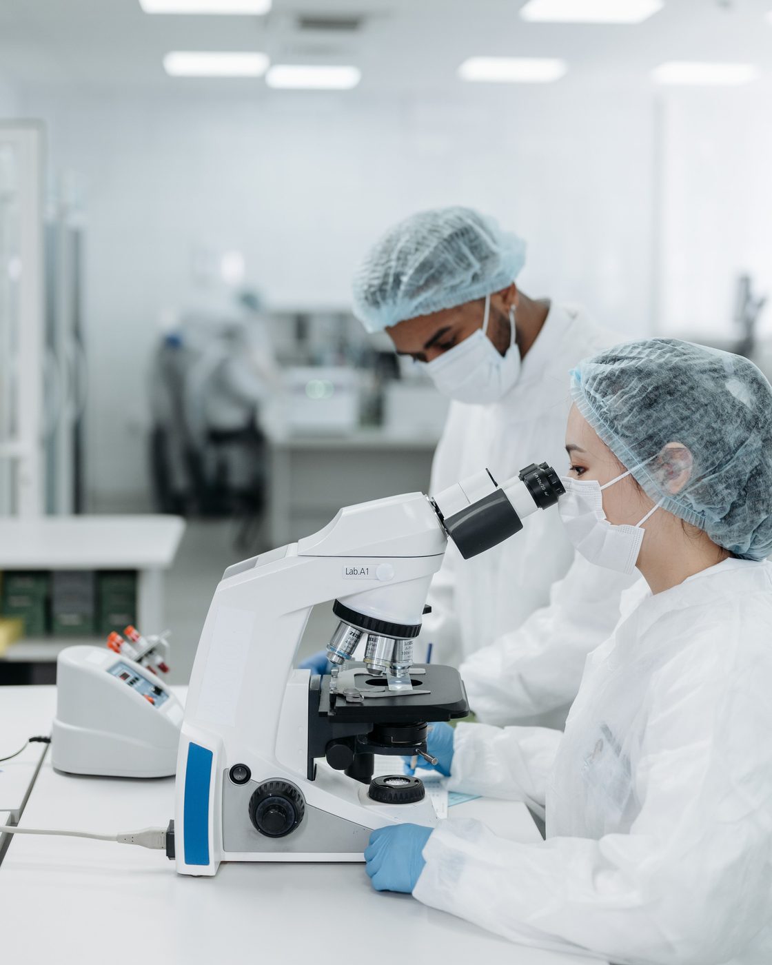 Two researchers in cleanroom gear at a laboratory bench, one looking through a microscope.